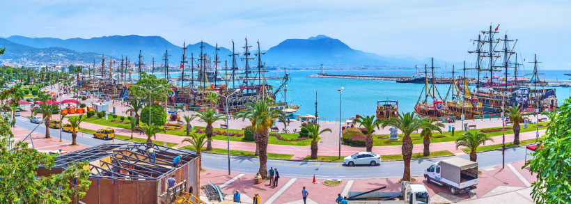 Blick auf den Hafen von Alanya mit mehreren bunt bemalten Ausflugsschiffen im Piratenstil, Palmenpromenade im Vordergrund und die imposanten Berge des Taurusgebirges im Hintergrund.