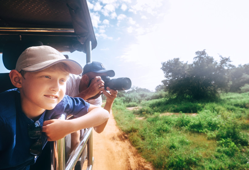 Ein Junge mit heller Kappe lehnt sich aus einem Safarifahrzeug und schaut neugierig in die Landschaft. Hinter ihm fotografiert ein Mann mit einer Kamera. Das Fahrzeug fährt auf einem sandigen Weg durch grünes Buschland unter leicht bewölktem Himmel. Die S
