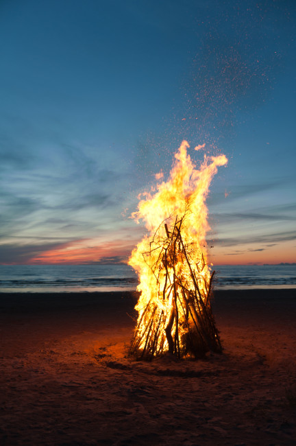 Lanzarote Großes Lagerfeuer aus aufgeschichteten Ästen brennt am Strand bei Sonnenuntergang. Der Himmel ist blau-orange gefärbt, das Meer liegt ruhig im Hintergrund.