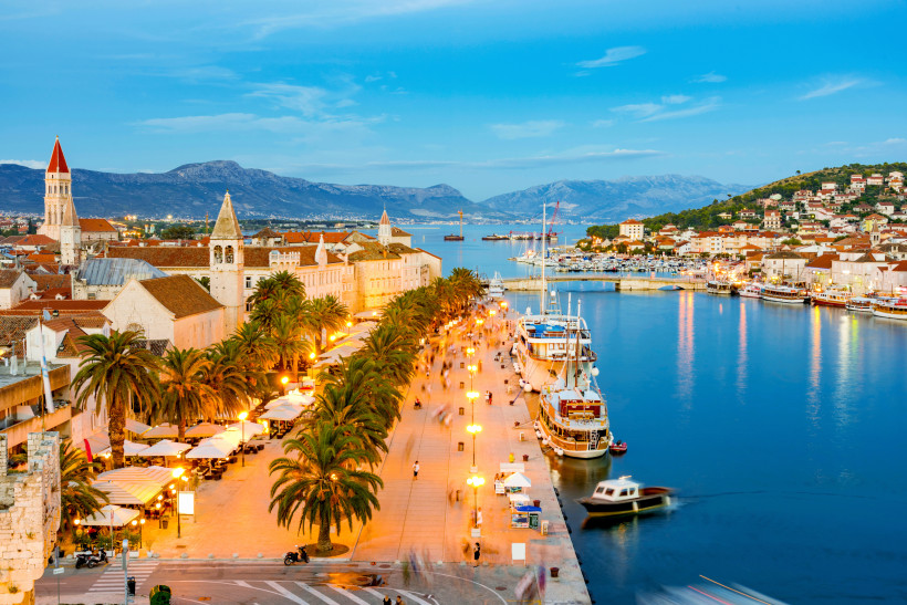 Abendstimmung an der Uferpromenade von Trogir in Kroatien. Entlang der beleuchteten Palmenallee flanieren Menschen, während Segelboote und Yachten im Hafen liegen. Im Hintergrund leuchten die historischen Gebäude und Kirchtürme der Altstadt, umrahmt von d