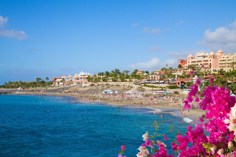 Sandstrand an der Küste mit Badegästen und Hotelanlage, im Vordergrund Bougainvillea Küstenansicht mit Sandstrand, Badegästen und Hotelanlage unter blauem Himmel, vorne pinke Bougainvillea