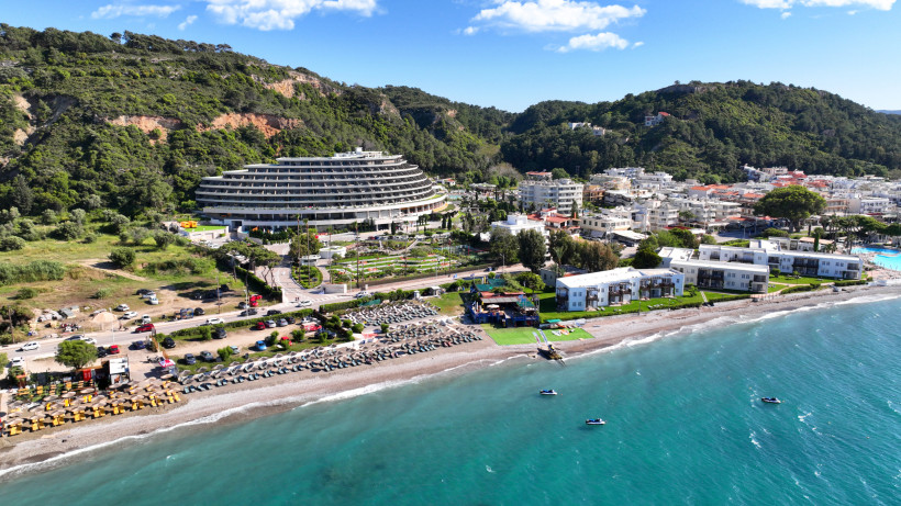 Luftaufnahme eines modernen Strandhotels an der Küste von Rhodos mit Poolanlage, Strand, türkisfarbenem Meer und grünen Hügeln im Hintergrund