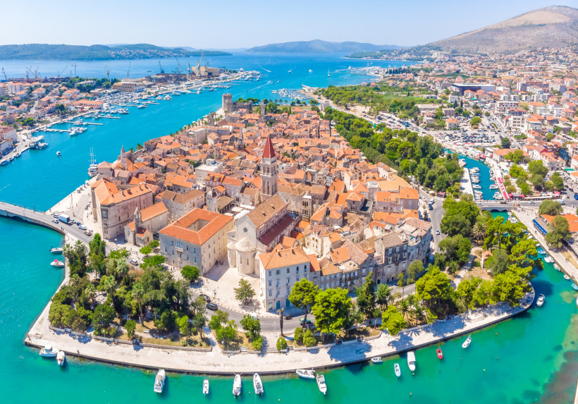 Luftaufnahme der Altstadt von Trogir in Kroatien. Die mittelalterliche Stadt liegt auf einer kleinen Insel, umgeben von türkisblauem Wasser. Zu sehen sind die historischen Gebäude mit roten Ziegeldächern, der markante Glockenturm der Kathedrale von Trogir