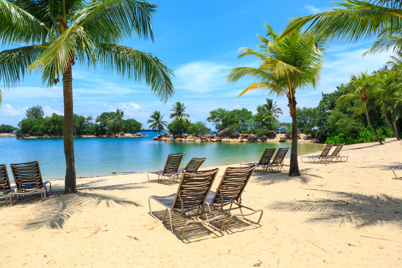 Singapur Mehrere leere Liegestühle stehen unter Palmen an einem ruhigen Sandstrand in Singapur. Im Hintergrund liegen kleine grüne Inseln in einer türkisblauen Bucht unter blauem Himmel mit weißen Wolken