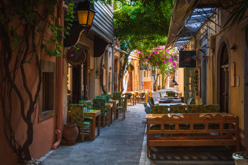 Schmale Gasse in der Altstadt von Rethymno mit Tischen und Stühlen vor Tavernen, überwachsen von Bougainvillea