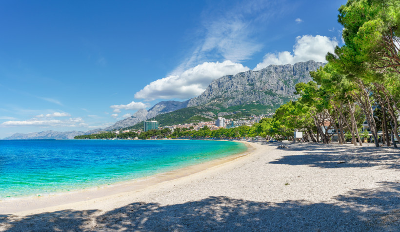 Kroatien Langer Kiesstrand mit türkisblauem Wasser, im Hintergrund eine Stadt vor imposanten, grauen Gebirgszügen. Am rechten Bildrand spenden Pinienbäume Schatten auf den Strand.