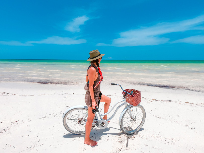 Junge Frau mit Fahrrad am weißen Sandstrand der Isla Holbox in Mexiko, türkisfarbenes Meer im Hintergrund – entspannter Karibikurlaub 2025/2026