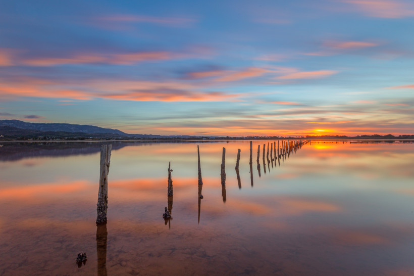 Kos Ruhige Salzlagune mit rosa und blauen Wolken im Wasser gespiegelt, Reihe alter Holzpfähle verläuft diagonal zum Sonnenuntergang