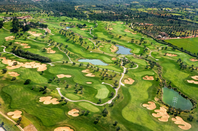 Mallorca Weitläufiger Golfplatz aus der Vogelperspektive mit saftig grünen Fairways, zahlreichen Sandbunkern und kleinen Teichen. Die gepflegte Anlage schlängelt sich durch eine mediterrane Landschaft mit Bäumen und umliegenden Hügeln. Im oberen Bereich sind Clubh