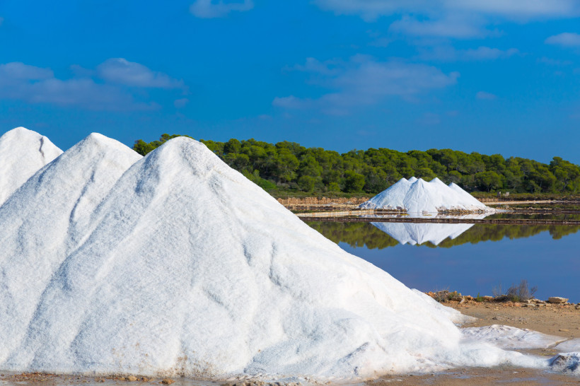 Mallorca - Ses Salines Salzberge in einer Saline auf Mallorca. Die Salzberge sind hoch und weiß, sehen aus wie kleine schneebedeckte Hügel, die sich vor einem klaren blauen Himmel und einem grünen Kiefernwald abheben. Im Vordergrund spiegelt sich der Himmel und die Salzberge in
