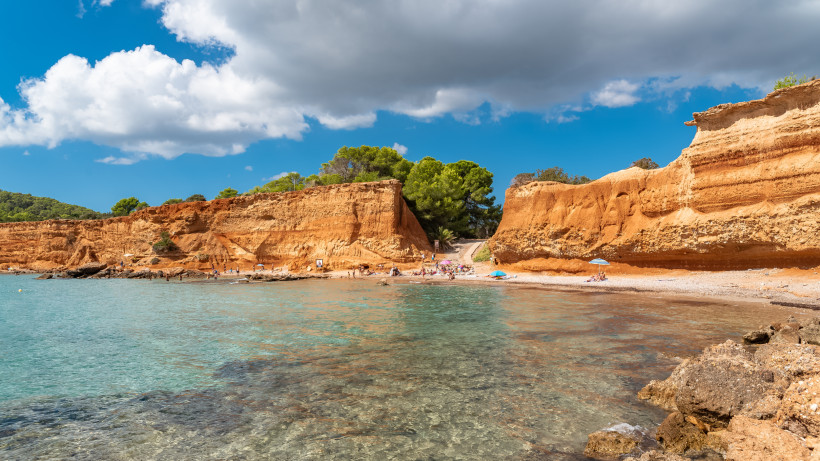 Bucht am Strand Sa Caleta auf Ibiza mit rotem Felskliff, klarem Wasser und vereinzelten Badegästen