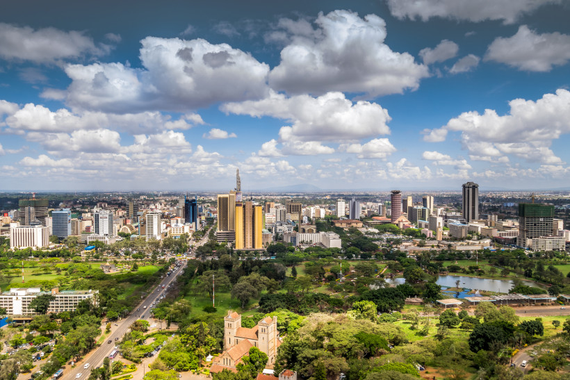 Kenia Panoramablick über Nairobi mit moderner Skyline, Hochhäusern, Grünflächen und Straßenverkehr. Weiße Wolken schweben über der Stadt, im Vordergrund ein Park mit Bäumen und einer Kirche