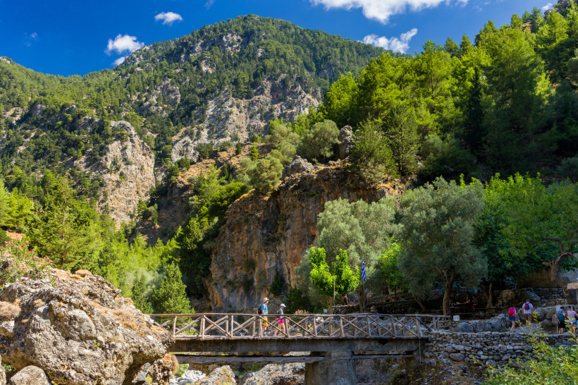 Wanderweg mit Holzbrücke in der Samaria-Schlucht vor bewaldeten Felsbergen unter blauem Himmel