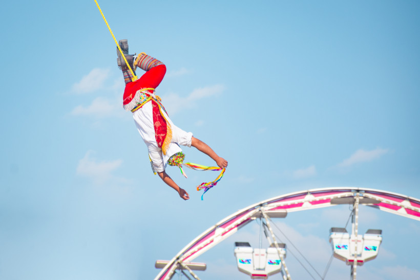 Volksfest in Aguascalientes Ein Volador de Papantla, ein traditioneller mexikanischer Lufttänzer, schwebt kopfüber an einem Seil, gekleidet in ein buntes Kostüm mit roten Hosen, weißen Oberteilen und bunten Bändern, mit einem Riesenrad im Hintergrund