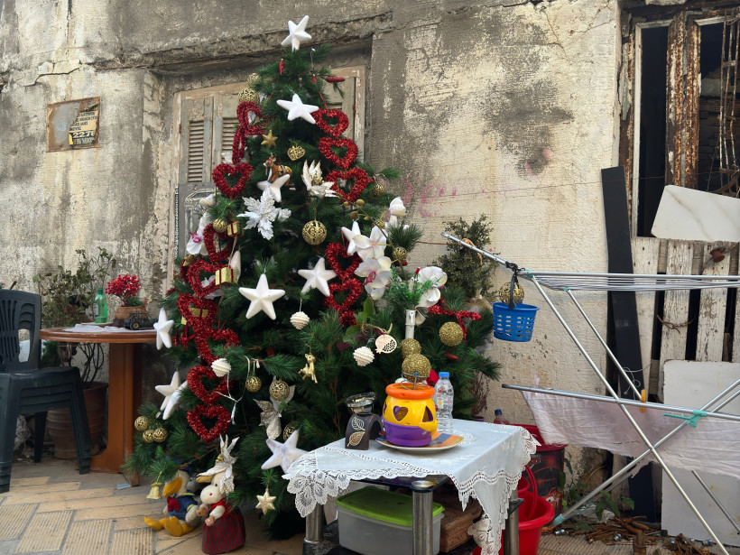 Kreta Ein liebevoll geschmückter Weihnachtsbaum mit roten Herzen, weißen Sternen und goldenen Kugeln steht vor einer verwitterten Hauswand in einer griechischen Gasse. Daneben ein kleiner Tisch mit einer Decke, Geschirr und Haushaltsgegenständen, sowie ein Wäsc
