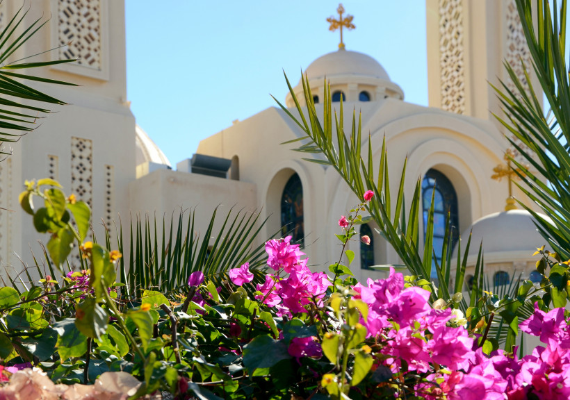 Ägypten - Sharm el Sheikh Große koptisch-orthodoxe Kirche mit mehreren runden Kuppeln, goldenen Kreuzen und einem markanten weißen Glockenturm. Die Fassade ist hellbeige und wirkt modern, mit großen Rundbogenfenstern und Glasmalereien. Der Himmel ist klar und blau.