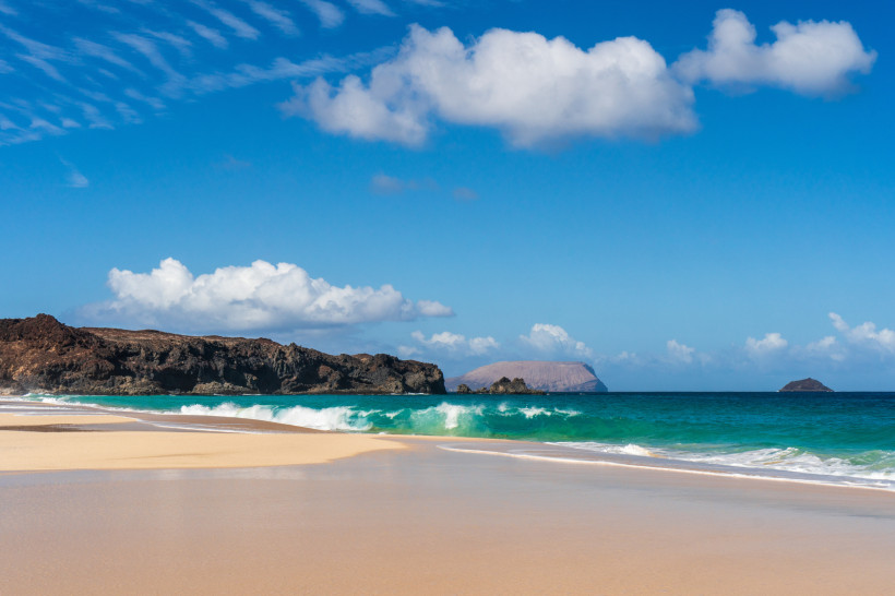 Playa de la Conchas mit hellem Sandstrand, türkisblauem Meer, Brandung und Felsküste unter blauem Himmel mit Wolken