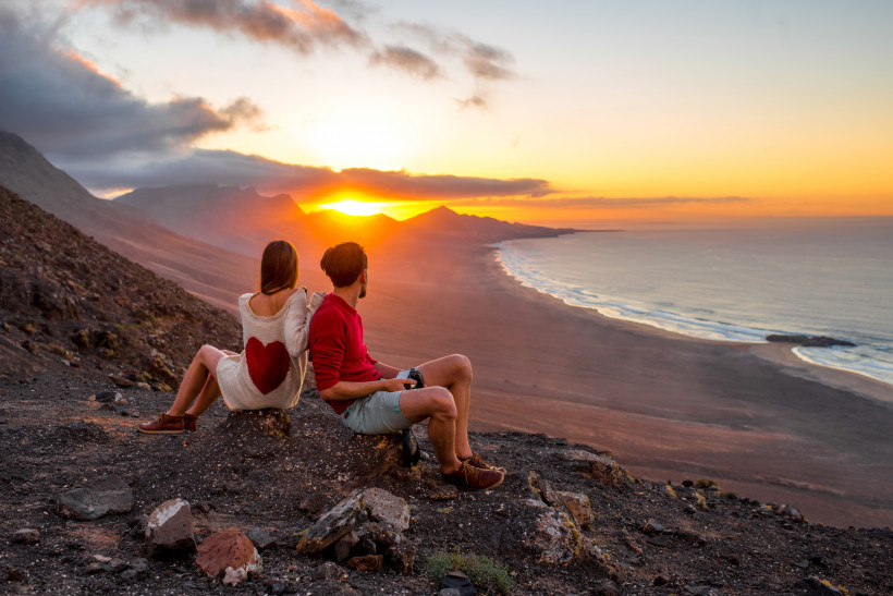 Fuerteventura Ein Paar sitzt auf einem Felsen und schaut in den Sonnenuntergang über einer weiten Küstenlandschaft mit Bergen und Meer. Die Frau trägt einen Pullover mit rotem Herz auf dem Rücken