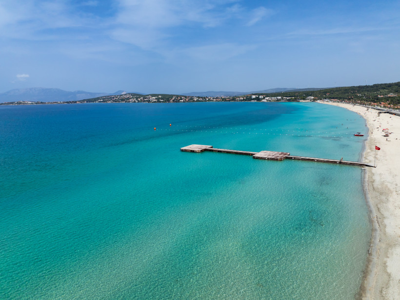 Panoramaaufnahme eines langen Sandstrands mit kristallklarem, türkisfarbenem Wasser an der türkischen Ägäis. Ein hölzerner Badesteg ragt ins Meer, im Hintergrund sind sanfte Hügel und Küstenorte zu sehen.