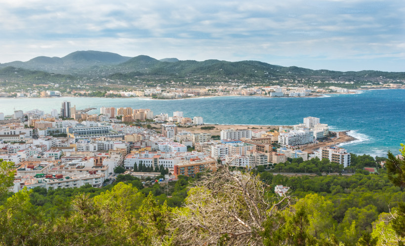 Sant Antoni de Portmany Blick auf die Küstenstadt Sant Antoni de Portmany auf Ibiza. Im Vordergrund grüne Pinienwälder, dahinter zahlreiche weiße und bunte Häuser bis hin zum Meer. Im Hintergrund die hügelige Landschaft der Insel.