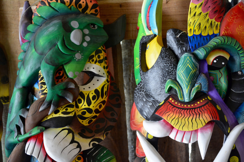 Juego de los Diablitos, Costa Rica Bunte, handgefertigte Holzmasken mit Tiermotiven, darunter ein grünes Leguan, ein Tukan und weitere fantasievolle, farbenfrohe Designs, ausgestellt an einer Holzwand