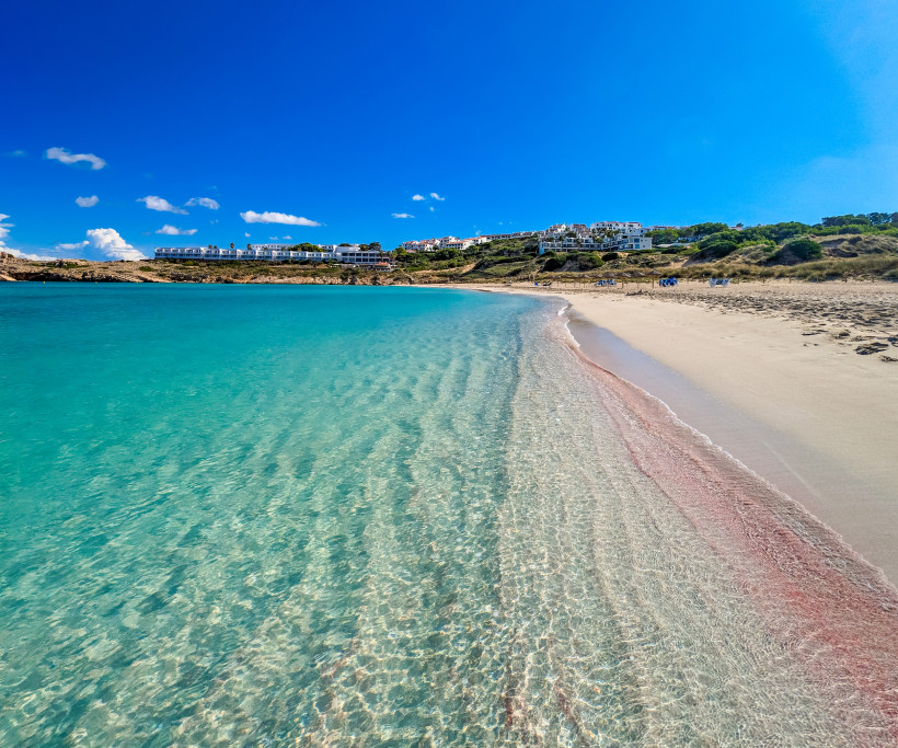 Flacher Sandstrand mit kristallklarem Wasser auf Menorca. Das Meer schimmert in türkis und verläuft sanft in den beigen Sand. Eine rötliche Algenlinie trennt den feuchten vom trockenen Sand. Im Hintergrund sind weiße Häuser und grüne Hügel zu sehen unter 