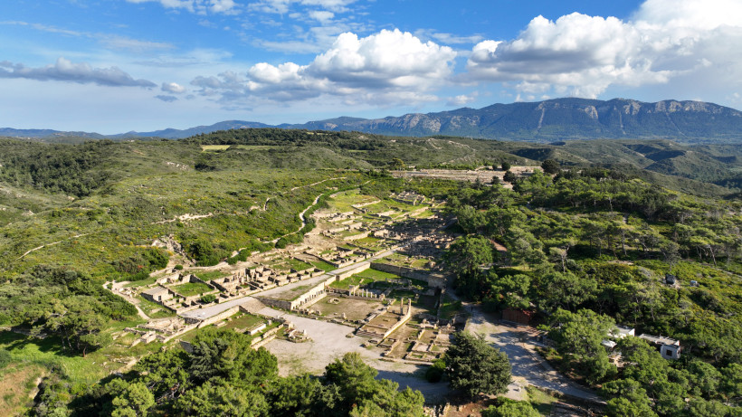Luftaufnahme einer weitläufigen antiken Ausgrabungsstätte mit Ruinen, Wegen und umgebender grüner Hügellandschaft vor einem Gebirge
