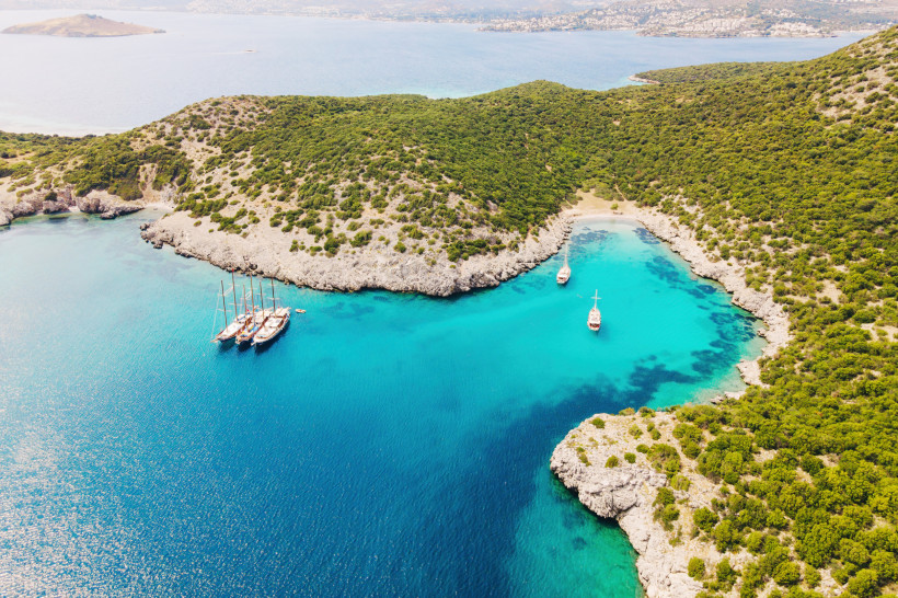 Luftaufnahme der Bucht am Akvaryum Beach mit türkisfarbenem Wasser, bewaldeten Hügeln und vor Anker liegenden Segelbooten