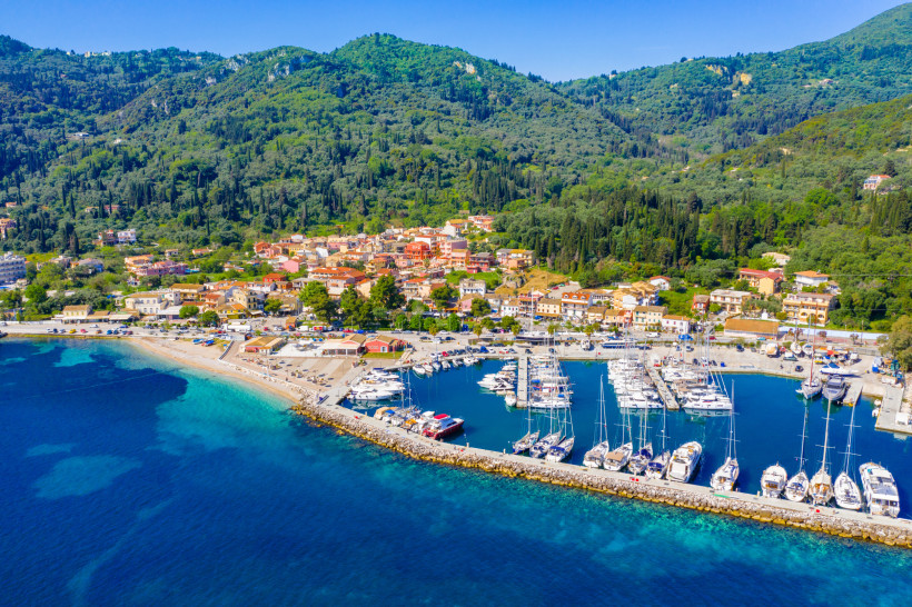 Es handelt sich um eine Luftaufnahme einer Küstenszene bei Tageslicht, die ein ruhiges blaues Meer zeigt, das ins tiefere Azurblau übergeht. Im Vordergrund sind vertäute Boote und ein Pier zu sehen. Ein Sandstrand mit Grün deutet auf einen beliebten Ort f
