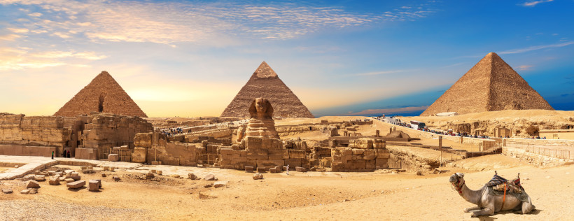 Ägypten Panoramablick auf die Pyramiden von Gizeh in Ägypten bei Sonnenuntergang. In der Mitte die berühmte Sphinx, im Vordergrund ein liegendes Kamel mit Sattel. Touristen sind auf dem Gelände unterwegs, der Himmel ist goldblau gefärbt.