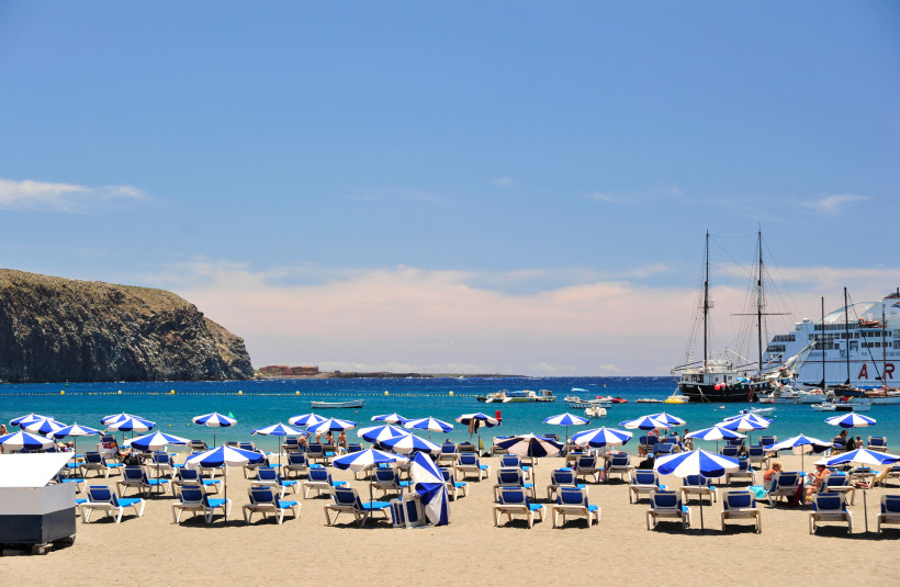 Strand mit Liegen und blau-weißen Sonnenschirmen in Arona auf Teneriffa, im Hintergrund Meer und Schiffe