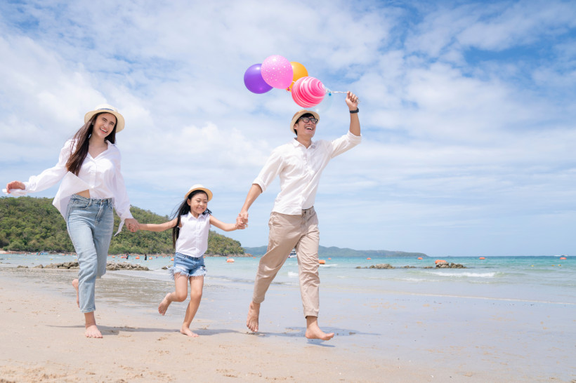 Thailand Pattaya Lachende Familie mit Kind läuft barfuß am Strand entlang. Der Vater hält bunte Luftballons, alle tragen helle Kleidung und Hüte. Im Hintergrund türkisblaues Meer und grüne Hügel