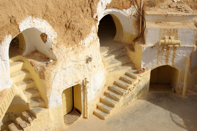 Höhlenwohnungen in Matmata, Tunesien. In den ockerfarbenen Felsen sind weiß gestrichene Türen und Fenster eingelassen. Kleine, verwinkelte Treppen führen zu den Eingängen, die in den Fels gehauen sind. Die Szene vermittelt ein Gefühl von Geschichte und tr