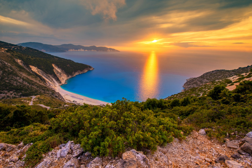 Blick auf Myrtos Beach mit türkisfarbener Bucht, hellem Strand und Sonnenuntergang über dem Meer