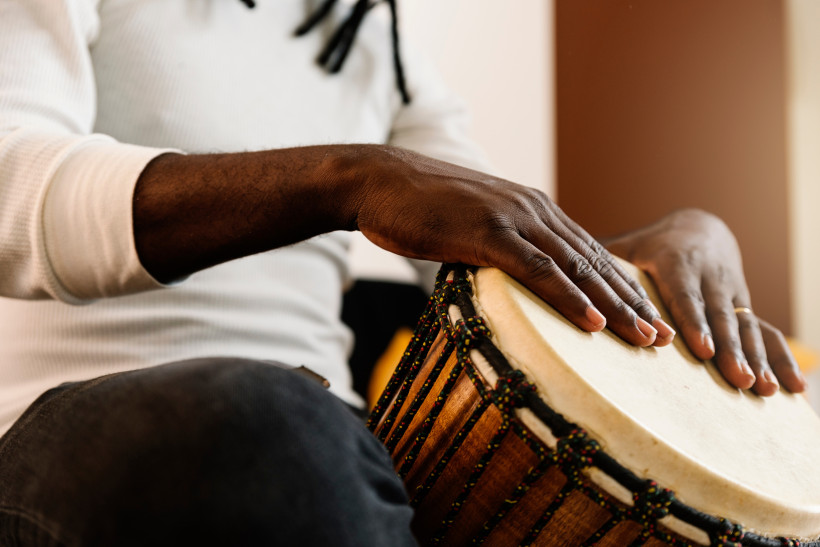 Jamaika Ein Mann spielt eine Djembe, eine traditionelle Trommel. Die Aufnahme zeigt den Oberkörper und die Hände, wie sie auf das Trommelfell schlagen. Der Mann trägt ein weißes Langarmshirt und sitzt mit überkreuzten Beinen. Die Djembe ist mit bunten Seilen verz