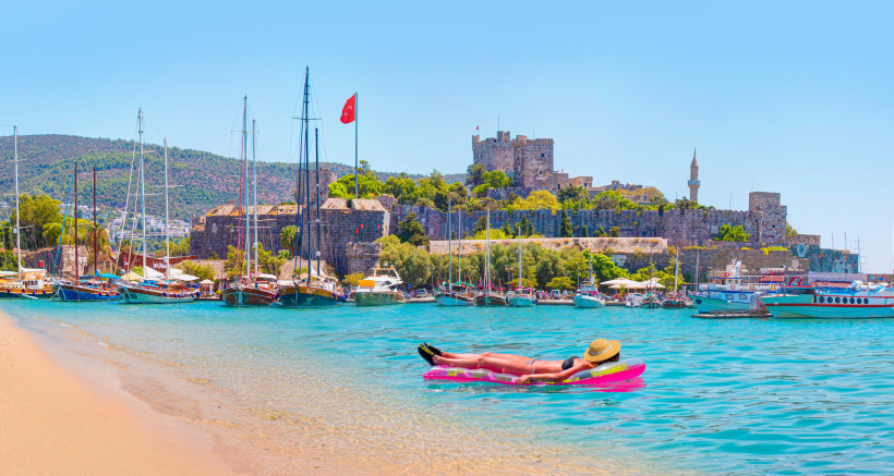 Entspannte Strandferien in Bodrum mit Person auf Luftmatratze im türkisblauen Meer, Yachthafen und historischer Burg an der türkischen Ägäis