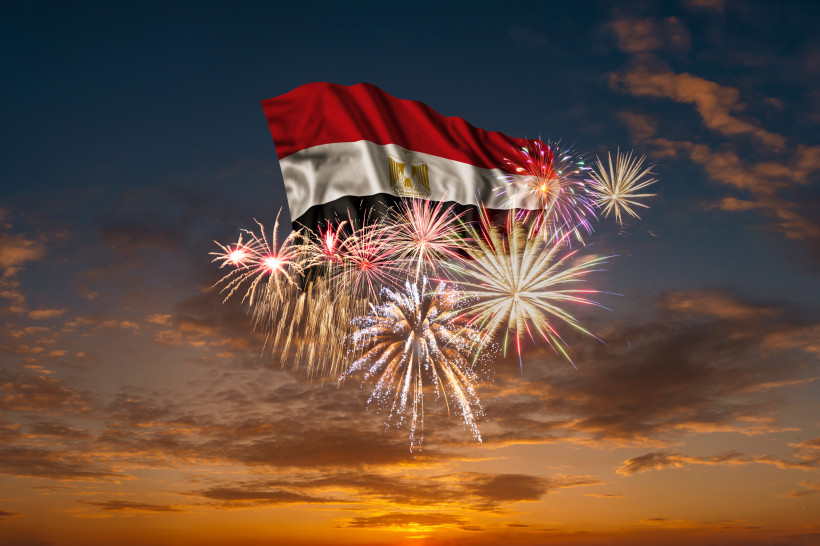 Ägypten - Sharm el Sheikh Ein spektakuläres Feuerwerk explodiert vor einem Sonnenuntergangshimmel. Darüber weht eine große ägyptische Flagge mit dem goldenen Adler in der Mitte.