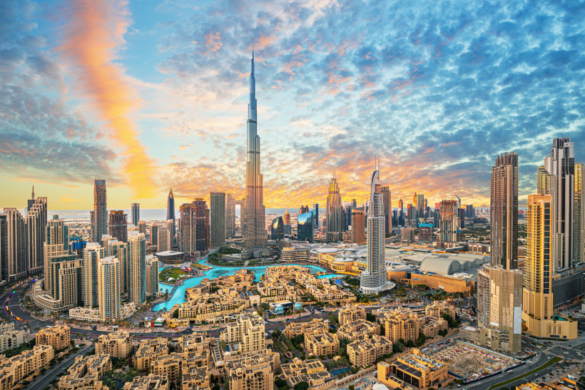 Panorama der Skyline von Downtown Dubai bei Sonnenuntergang. Im Zentrum steht der Burj Khalifa, umgeben von modernen Hochhäusern, Wasserläufen und Hotels. Der Himmel zeigt eine dramatische Wolkenformation mit orange-blauen Lichtverläufen. Im Vordergrund s