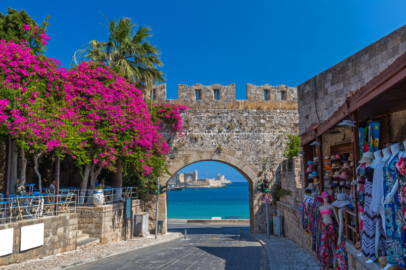 Historisches Stadttor in der Altstadt von Rhodos mit blühender Bougainvillea, Palmen und Blick auf das türkisfarbene Meer
