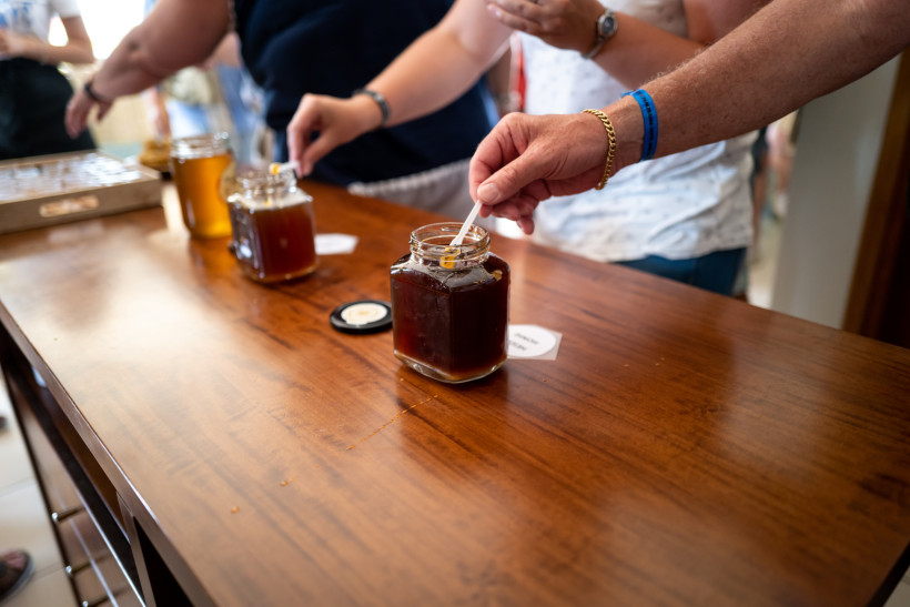 Mehrere Personen stehen an einem Holztresen und probieren Honig aus offenen Gläsern. Einer der Gäste taucht einen kleinen Probierlöffel in ein Glas mit dunklem Honig. Im Hintergrund sind weitere Honiggläser, Deckel und Menschen zu sehen.