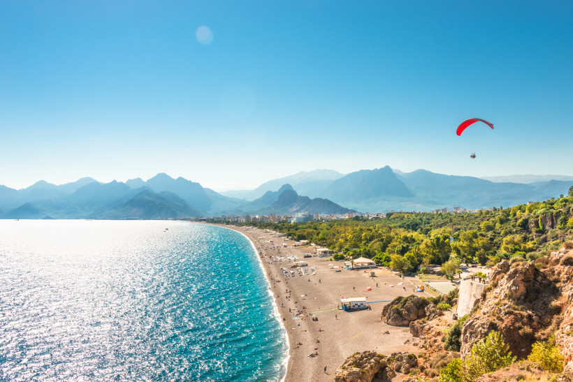 Weitläufiger Konyaaltı Strand in Antalya mit glasklarem Wasser, Bergkulisse und Paraglider – beliebtes Highlight im Antalya Urlaub.
