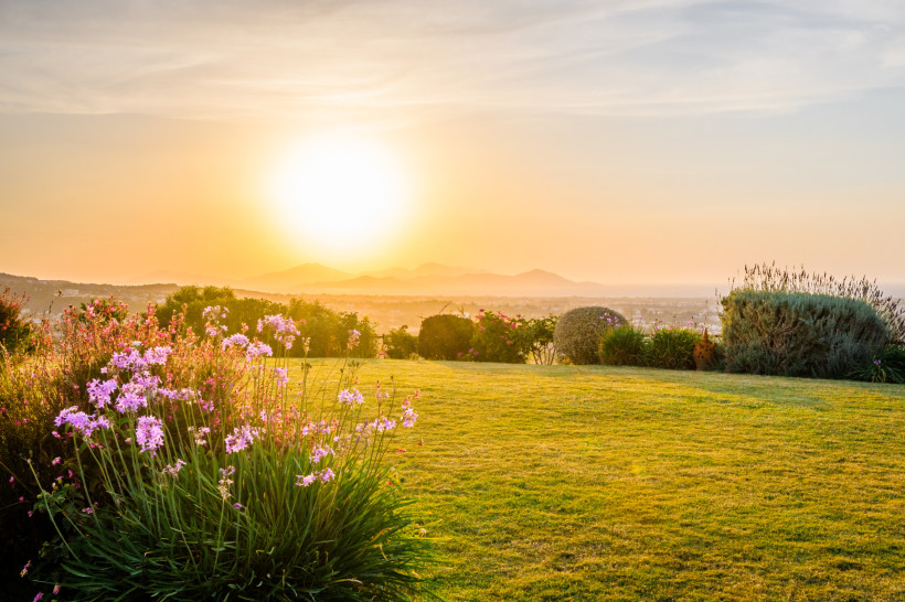 Ein weitläufiger Garten mit gepflegtem Rasen und blühenden Blumen, aufgenommen im warmen Licht der untergehenden Sonne. Im Hintergrund ist die Silhouette eines Hügellands mit sanften Bergketten zu erkennen. Der Himmel leuchtet in sanften Orange- und Gelbt