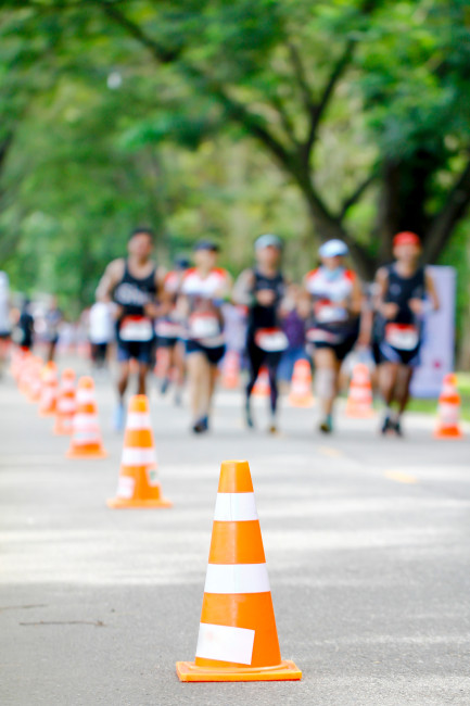 Formentera Laufveranstaltung auf einer von Bäumen gesäumten Straße, im Vordergrund orange-weiße Verkehrskegel, im Hintergrund Läuferinnen und Läufer in Sportkleidung, teils unscharf.