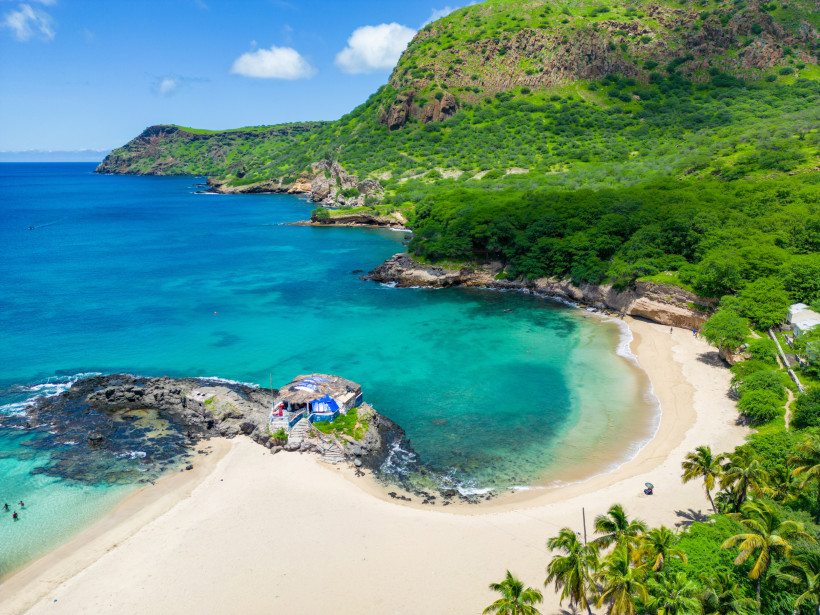 Luftaufnahme einer traumhaften Bucht auf den Kapverden mit grün bewaldeten Hügeln, steilen Felsen und klarem, türkisfarbenem Wasser. In der Mitte eine kleine Felseninsel mit einer Strandbar. Der helle Sandstrand ist von Palmen gesäumt.