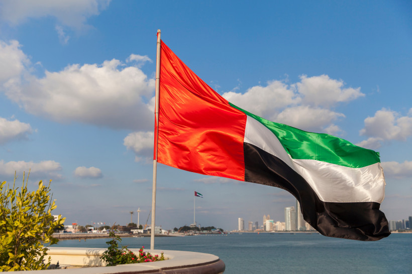 Die Flagge der Vereinigten Arabischen Emirate weht im Wind mit einer Stadtlandschaft und einem Wasserblick im Hintergrund