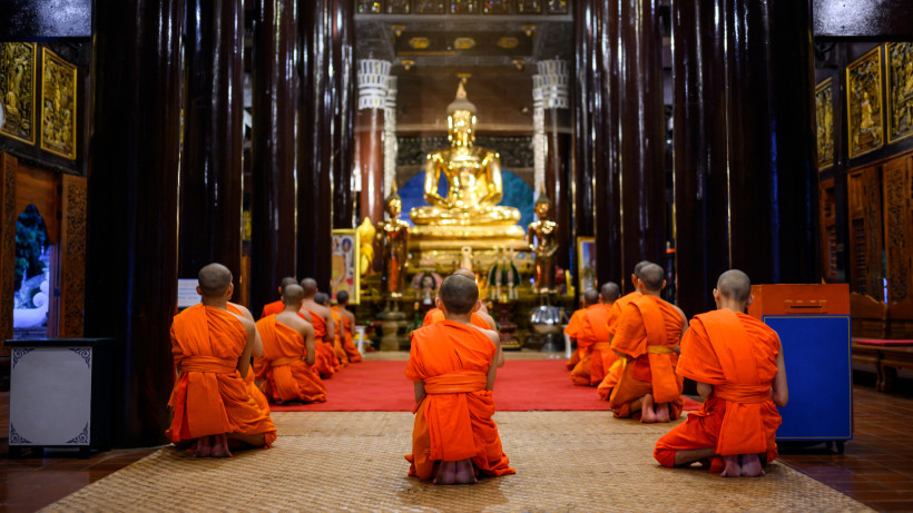 Thailand Pattaya Gruppe buddhistischer Mönche in oranger Robe kniet in einem Tempel vor einer großen goldenen Buddha-Statue. Die Atmosphäre wirkt ruhig und ehrfürchtig, die Halle ist mit dunklen Holzsäulen und goldenen Verzierungen geschmückt.