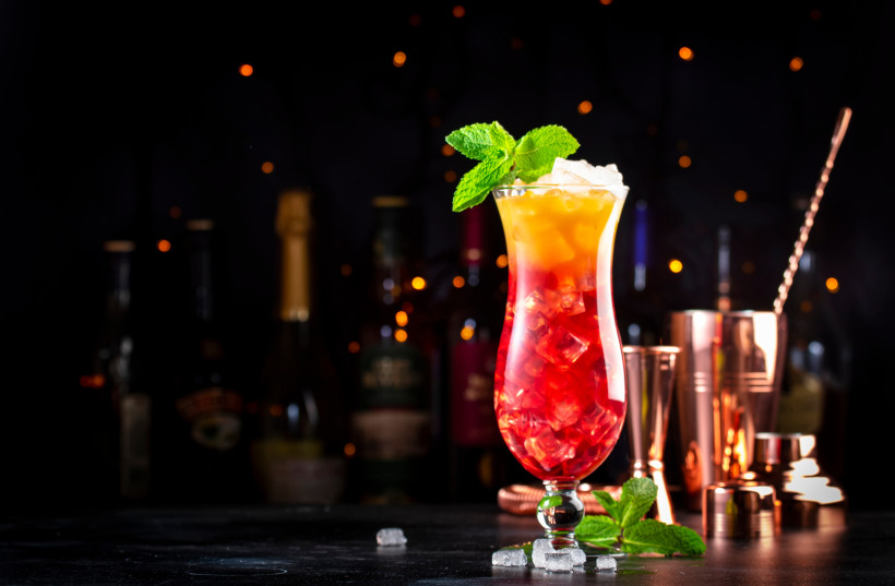 Leuchtend bunter Cocktail in einem hohen Glas, serviert mit Eiswürfeln und Minze. Im Hintergrund stehen Flaschen und Barkeeper-Equipment, umrahmt von stimmungsvollen Lichtern auf dunklem Hintergrund.