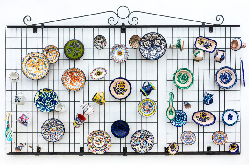 Fuerteventura Bunte, handbemalte Keramikteller, Tassen und Schalen hängen dekorativ an einem Gitter vor weißer Wand. Die Muster sind vielfältig, meist floral oder geometrisch
