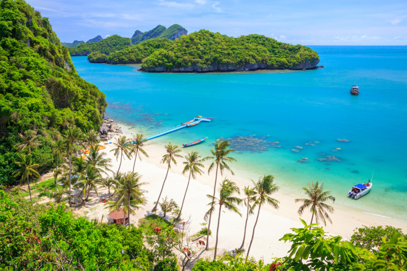 Thailand - Koh Samui Das Bild zeigt eine traumhafte tropische Bucht mit weißem Sandstrand, der von hohen Palmen gesäumt ist. Das türkisfarbene Wasser ist kristallklar und geht fließend in ein tiefes Blau über. Am Ufer liegen mehrere bunte Longtailboote sowie ein moderneres Mo