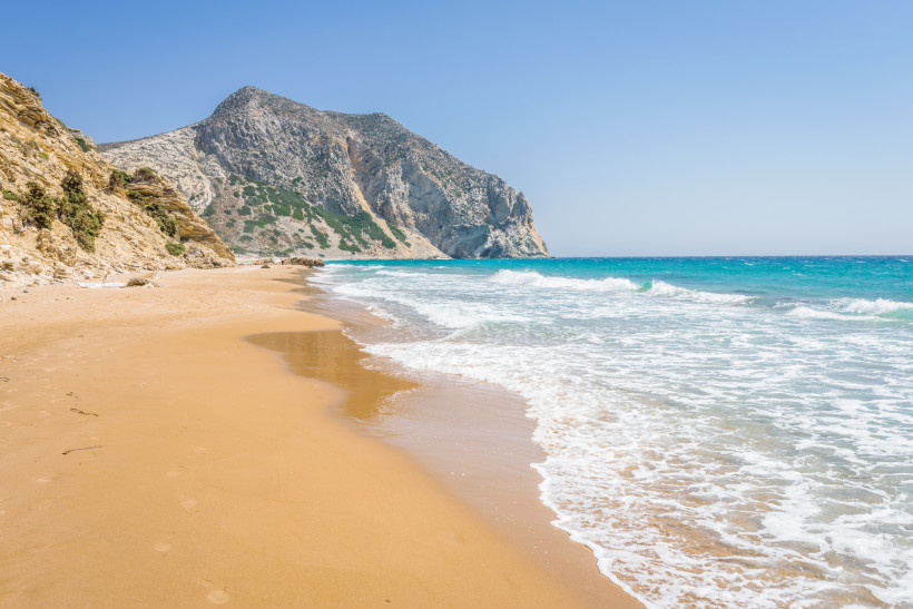 Weitläufiger, menschenleerer Sandstrand mit goldfarbenem Sand und sanften Wellen, die an das Ufer rollen. Im Hintergrund ragt ein steiler, felsiger Berg empor. Die Landschaft wirkt ursprünglich und naturbelassen, das Wasser schimmert türkis und der Himmel